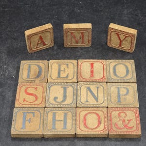 May include: A collection of fifteen square wooden blocks featuring embossed letters and symbols. Three blocks are arranged in a row above a grid of twelve blocks. The letters A, M, Y, D, E, I, O, S, J, N, P, F, H, O, and the ampersand symbol are printed in red or blue.