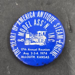 May include: A round blue pinback button featuring a white graphic of a steam engine tractor. The text reads: THE HEART OF AMERICA ANTIQUE STEAM ENGINE & MODEL ASS'N. INC. 17th Annual Reunion Aug. 2-3-4, 1974 MCLOUTH, KANSAS in white lettering.
