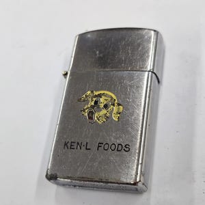 May include: A silver-toned metal lighter with a hinged lid. The lighter features a yellow and black cartoon dog graphic and the text "KEN-L FOODS" printed on the front. The lighter shows signs of wear.