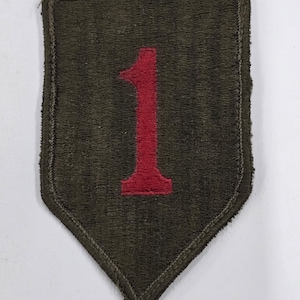May include: A dark green, shield-shaped patch with a red numeral "1" embroidered in the center. The patch has a stitched border and a pointed bottom. The patch is likely a military insignia or emblem.