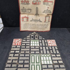 May include: A set of vintage architectural building blocks arranged to form a house facade, accompanied by a paper instruction sheet titled The Embossing Company's Architectural Blocks No. 101, featuring diagrams of various structures to build.