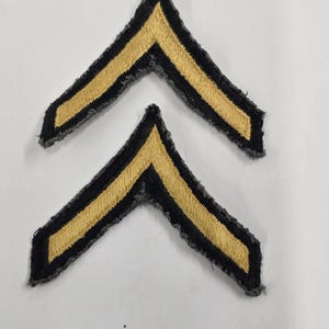 May include: Two chevron-shaped embroidered patches featuring a central gold-colored stripe bordered by black fabric edges. The patches have a distinct V-shape design with frayed, textured borders, showcasing a classic military-style insignia aesthetic.