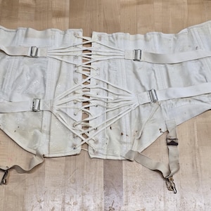 May include: A white corset featuring a central lace-up closure with white cord, adjustable side straps with silver-toned metal buckles, and four attached garter straps with metal clips. The garment has a textured fabric finish throughout.