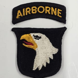 May include: A black embroidered patch featuring the word AIRBORNE in yellow text, paired with a shield-shaped patch displaying a white eagle head with a yellow beak and red tongue on a black background, both with black stitched borders.
