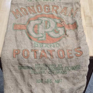 May include: A vintage burlap potato sack with the words "MONOGRAM" and "POTATOES" printed in orange and green. The sack is made of a coarse, woven material and shows signs of wear.