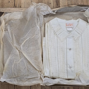 May include: A white button-down shirt with a pointed collar and vertical lace detailing, presented inside a cardboard box lined with crinkled tissue paper. The shirt features a label reading "Saks Fifth Avenue" at the inner neckline.