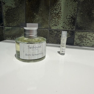 Pode incluir: Um frasco de perfume redondo de vidro com tampa prateada e um rótulo com a inscrição Toskovat Born Screaming, acompanhado por um pequeno frasco de amostra transparente com um rótulo contendo o texto Etsy e ebay, ambos com fragrância amarela.