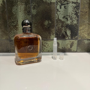 Puede incluir: Un frasco de perfume rectangular de vidrio con tapón plateado y un frasco pulverizador transparente. El perfume del interior es de color ámbar. La botella tiene la palabra "YOU" en la parte delantera. El fondo es una pared texturizada de color verde.