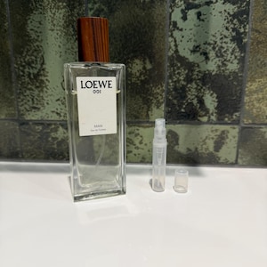 Puede incluir: Una botella rectangular de vidrio de Loewe 001 Man Eau de Toilette con tapa de madera. La botella tiene una etiqueta blanca con texto negro. Un pequeño frasco pulverizador transparente y una tapa están junto a la botella de perfume.
