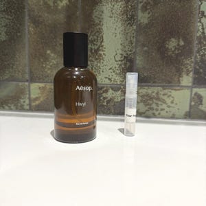 May include: A brown glass bottle of Aesop Hwyl Eau de Parfum with a black cap. A small clear vial with the text "Your frag" is next to the bottle. The bottle has a brown and orange striped design.