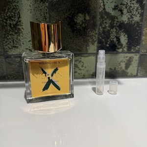 May include: A clear glass perfume bottle with a gold cap and label. The label reads "Wulong Cha X" and "Nishane Extrait de Parfum Shanghai". A small, clear spray bottle and cap are next to the perfume bottle.
