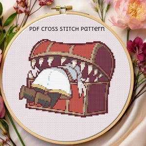 Pode incluir: Padrão de ponto cruz em um bastidor de madeira. O design apresenta um baú do tesouro vermelho e marrom com dentes afiados, aberto para revelar um objeto branco. O texto "PDF CROSS STITCH PATTERN" está no topo. O fundo inclui flores cor-de-rosa.