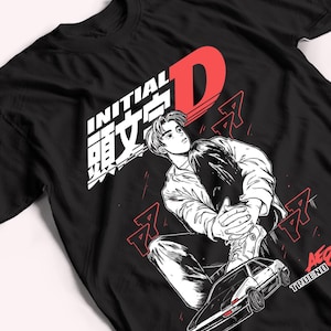 Puede incluir: Una camiseta negra con un estampado gráfico de un personaje sentado sobre un coche. El diseño incluye el texto Initial D en blanco y rojo, caracteres japoneses y el texto AE86 TRUENO. La ilustración utiliza tinta negra, blanca y roja.