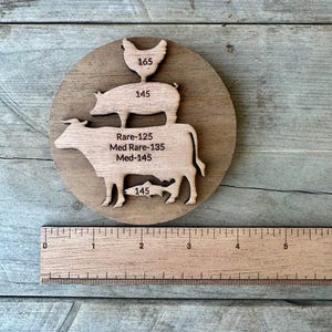 May include: Wooden cutting board with a circular design featuring stacked animal cutouts: a chicken, pig, cow, and fish. The board includes temperature guides for cooking meat. A wooden ruler is placed below the board.