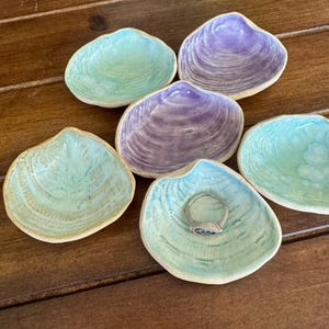 May include: Six ceramic seashell-shaped dishes in shades of teal and purple. One dish holds a silver ring with a blue stone. The dishes have a textured, glazed finish and are arranged on a wooden surface.