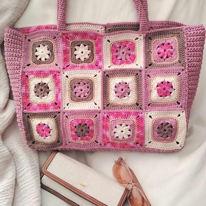 Peut inclure: Un sac fourre-tout crocheté à la main dans les tons de rose, marron et crème, avec un motif floral carré. Le sac a une poignée rose assortie, un portefeuille beige et des lunettes de soleil roses.