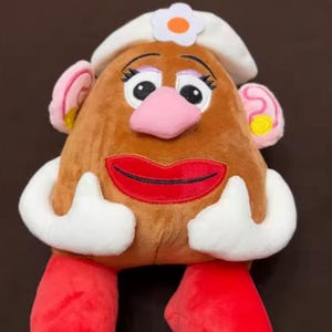 May include: A plush toy resembling a potato with a brown body, pink nose, and red lips. It wears a white hat with a flower and has pink ears with yellow accents. The toy has white arms and red feet.