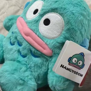 May include: A plush turquoise Hangyodon character with large white eyes, a pink tongue, and blue fins. The plush toy has a tag with the Hangyodon logo and name. The toy is on a soft, light-colored surface.
