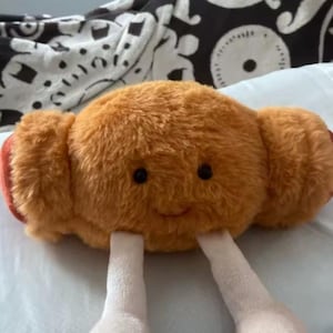 May include: A soft toy croissant with a smiling face, black eyes, and two white legs. The croissant is light brown with red ends. The toy is fluffy and appears to be made of a soft material.