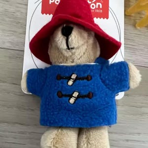 May include: A Paddington Bear plush keychain. The bear wears a red hat and a blue coat with toggle closures. The plush toy has a blue loop for attaching to keys or a bag. The tag reads "Paddington" and "18m+".