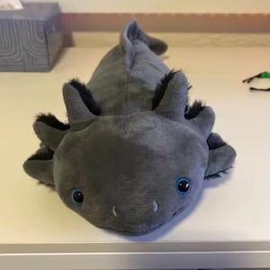 Pode incluir: Um brinquedo de pelúcia cinza de axolotl com detalhes em preto ao redor das guelras e da cabeça. O brinquedo tem olhos azuis e está sobre uma superfície branca. O fundo inclui uma caixa de lenços e uma parede com bolinhas vermelhas.