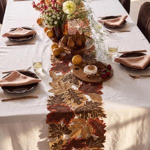 Puede incluir: Un camino de mesa con una disposición superpuesta de hojas de tela en tonos marrón, naranja y dorado. El camino se coloca sobre un mantel blanco con platos oscuros, servilletas rosas, cubiertos de cobre, un centro floral y frutas.