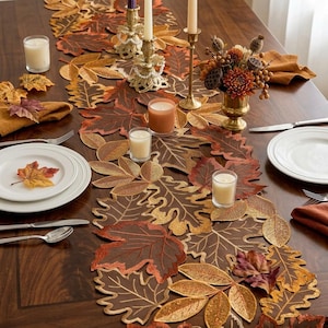 Può includere: Un runner da tavola con un motivo di foglie autunnali ricamate nei toni dell'arancione, marrone e oro. Il runner è decorato con due candelabri dorati, quattro candele votive in vetro e un centrotavola floreale su un tavolo in legno.