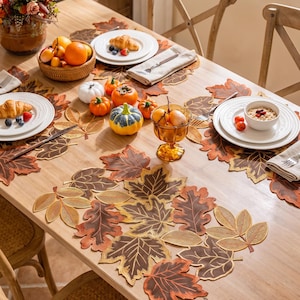 May include: A set of brown, orange, and gold leaf-shaped placemats and a table runner featuring intricate embroidered vein details. The table setting includes white plates, linen napkins, small decorative pumpkins, and a glass bowl of fruit.