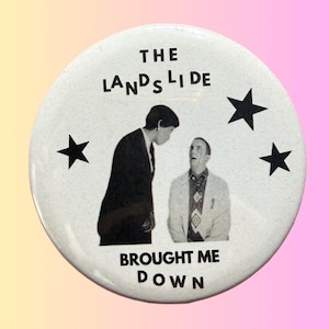 Peut inclure: Un bouton blanc avec le texte "THE LANDSLIDE BROUGHT ME DOWN" et une image en noir et blanc de deux hommes. Trois étoiles noires sont également présentes. Le bouton a une finition brillante.