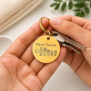 Puede incluir: Un llavero redondo de metal dorado con una ilustración grabada de una familia de figuras de palo, un corazón, una flor y el texto "Meine Familie" en negro. El dije circular está unido a una anilla de metal dorado que sostiene dos llaves plateadas.