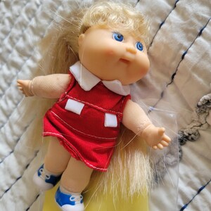 K&ouml;nnte beinhalten: Eine Vintage-Puppe mit blonden Haaren und blauen Augen. Die Puppe tr&auml;gt ein rotes Kleid mit wei&szlig;em Kragen und wei&szlig;en Details sowie blaue und wei&szlig;e Turnschuhe. Ein klassisches Spielzeug.