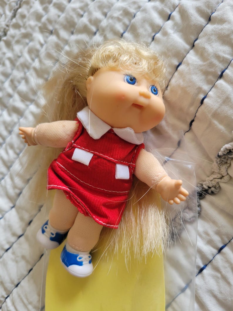 K&ouml;nnte beinhalten: Eine Vintage-Puppe mit blonden Haaren und blauen Augen. Die Puppe tr&auml;gt ein rotes Kleid mit wei&szlig;em Kragen und wei&szlig;en Details sowie blaue und wei&szlig;e Turnschuhe. Ein klassisches Spielzeug.