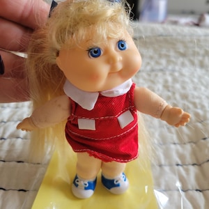 K&ouml;nnte beinhalten: Eine kleine Puppe mit blonden Haaren, blauen Augen und einem roten Kleid mit wei&szlig;en Akzenten. Die Puppe tr&auml;gt blaue und wei&szlig;e Turnschuhe. Die Arme der Puppe sind ausgestreckt, und sie steht auf einer gelben Oberfl&auml;che.