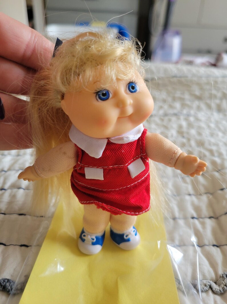 K&ouml;nnte beinhalten: Eine kleine Puppe mit blonden Haaren, blauen Augen und einem roten Kleid mit wei&szlig;en Akzenten. Die Puppe tr&auml;gt blaue und wei&szlig;e Turnschuhe. Die Arme der Puppe sind ausgestreckt, und sie steht auf einer gelben Oberfl&auml;che.