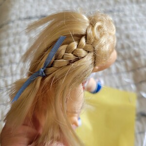 K&ouml;nnte beinhalten: Blondes Puppenhaar mit einem seitlichen Zopf und einem blauen Band. Das Haar der Puppe ist hellblond und der Zopf ist ordentlich gemacht. Das blaue Band f&uuml;gt der Frisur einen Farbtupfer hinzu. Der Arm der Puppe ist sichtbar.