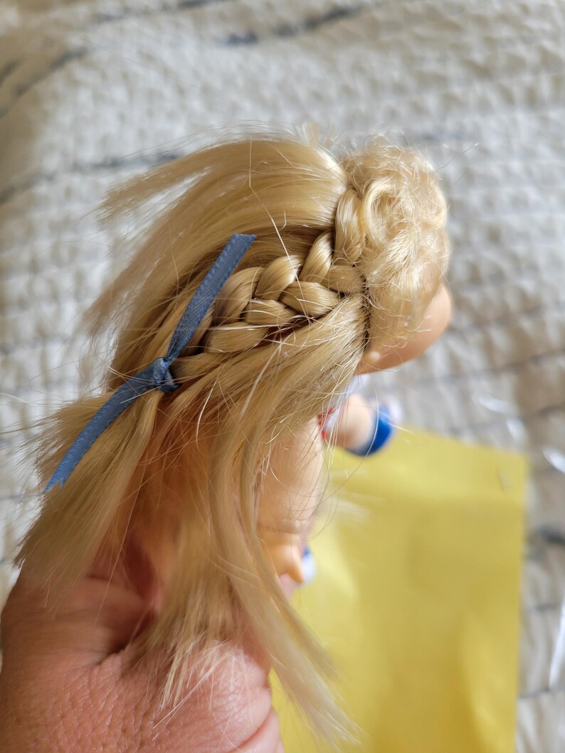 K&ouml;nnte beinhalten: Blondes Puppenhaar mit einem seitlichen Zopf und einem blauen Band. Das Haar der Puppe ist hellblond und der Zopf ist ordentlich gemacht. Das blaue Band f&uuml;gt der Frisur einen Farbtupfer hinzu. Der Arm der Puppe ist sichtbar.