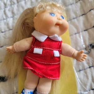May include: A vintage doll with blonde hair in a ponytail, blue eyes, and rosy cheeks. The doll wears a red dress with a white collar and white accents, paired with blue and white sneakers. The doll is a collectible item.