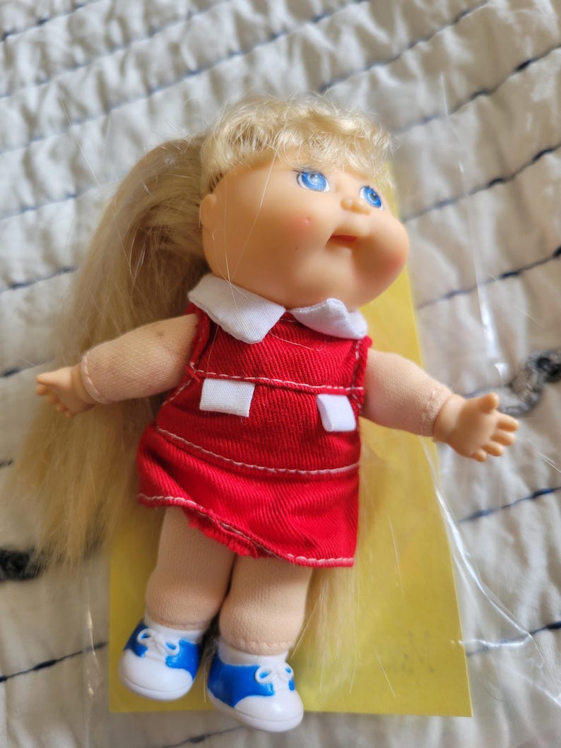 K&ouml;nnte beinhalten: Eine Vintage-Puppe mit blonden Haaren im Pferdeschwanz, blauen Augen und rosigen Wangen. Die Puppe tr&auml;gt ein rotes Kleid mit wei&szlig;em Kragen und wei&szlig;en Akzenten, kombiniert mit blau-wei&szlig;en Turnschuhen. Ein Sammlerst&uuml;ck.