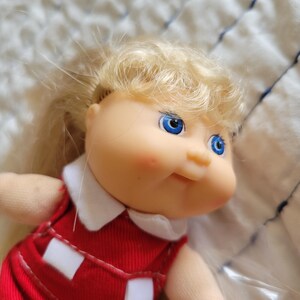 K&ouml;nnte beinhalten: Eine Vintage-Puppe mit blondem Haar und blauen Augen. Die Puppe tr&auml;gt ein rotes Kleid mit wei&szlig;en Akzenten und einem wei&szlig;en Kragen. Das Gesicht der Puppe hat rosige Wangen und ein leichtes L&auml;cheln. Eine Sammlerpuppe.
