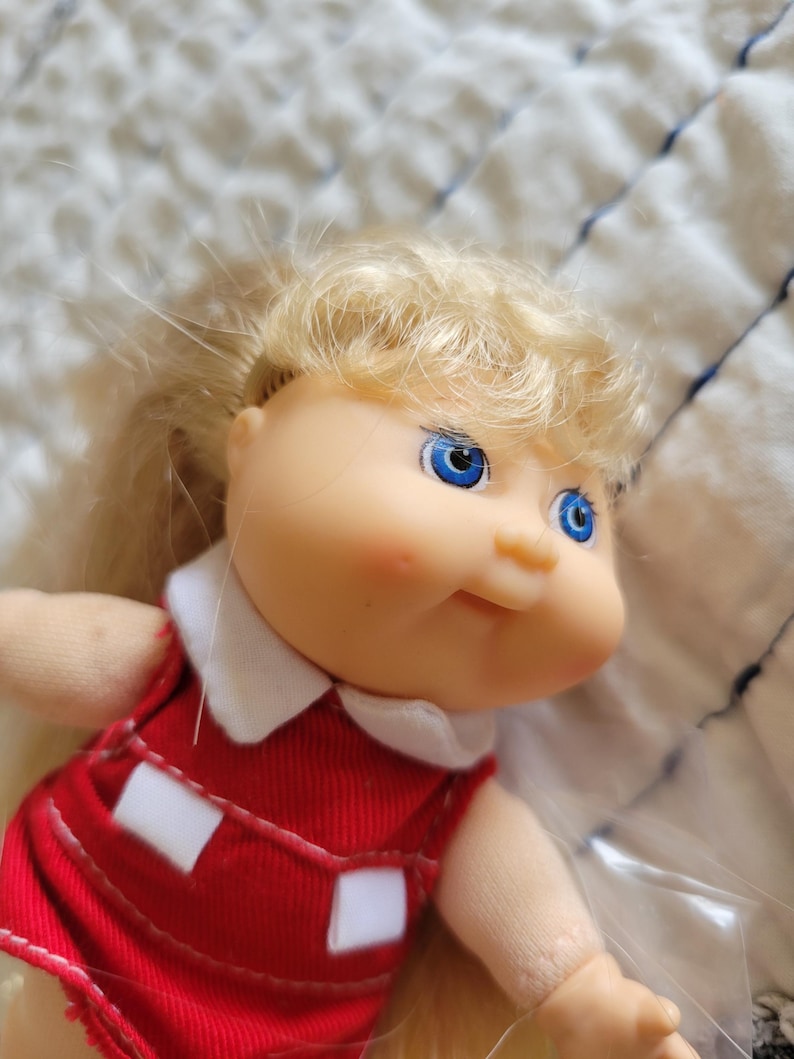 K&ouml;nnte beinhalten: Eine Vintage-Puppe mit blondem Haar und blauen Augen. Die Puppe tr&auml;gt ein rotes Kleid mit wei&szlig;en Akzenten und einem wei&szlig;en Kragen. Das Gesicht der Puppe hat rosige Wangen und ein leichtes L&auml;cheln. Eine Sammlerpuppe.