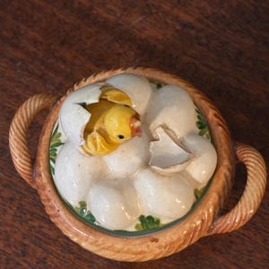 May include: A ceramic basket-shaped container with a lid featuring a chick hatching from an egg. The chick is yellow, and the eggs are white. The basket has a woven texture and brown handles. Green leaves are painted around the eggs.