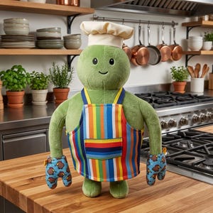 May include: A green plush toy in a chef's hat and apron. The apron has colorful vertical stripes. The toy wears oven mitts with a blue background and turkey pattern. It stands on a wooden countertop in a kitchen setting.