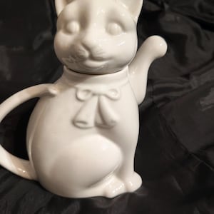Może przedstawiać: Biały ceramiczny czajnik w kształcie kota, z kocią twarzą jako pokrywką i wzorem kokardy. Czajnik ma uchwyt i dzióbek, na ciemnym tle.