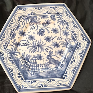 Puede incluir: Plato hexagonal de cerámica con diseño floral azul y blanco. El plato presenta un borde con un patrón repetitivo y un diseño central con flores, follaje y un conejo. El plato tiene un fondo crema claro.