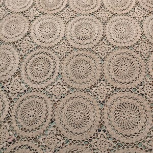 Pode incluir: Close-up de um têxtil bege de crochê. O design apresenta um padrão repetido de grandes toalhas de mesa circulares, conectadas por um design de trabalho aberto. Isso cria uma superfície texturizada com uma aparência vintage e artesanal.