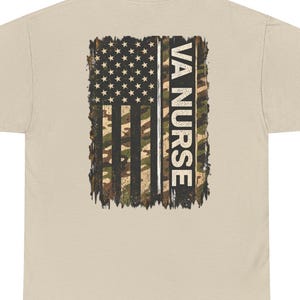 May include: A sand-coloured t-shirt featuring a graphic on the back. The design displays a distressed American flag pattern with a camouflage print and the vertical text VA NURSE in white capital letters, set against a dark, textured background.