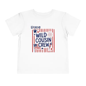 Puede incluir: Una camiseta blanca de manga corta con un estampado gráfico que incluye el texto Dixie WILD COUSIN CREW en azul. El diseño presenta rayas verticales rojas, una pequeña bandera estadounidense, estrellas azules, un fuego artificial y un cohete.