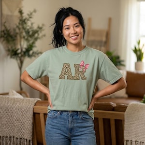 May include: A pale green t-shirt featuring the letters "AR" in a leopard print pattern, adorned with a pink bow. The shirt is worn with blue denim jeans.