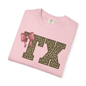 Op de afbeelding: Lichtroze T-shirt met de letters "TX" in een luipaardprint en een gouden rand. Een roze strik met een strass-middenstuk bevindt zich aan de linkerkant van de "T". Het shirt is opgevouwen en heeft een label in de rechterbovenhoek.