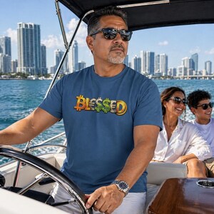 Puede incluir: Una camiseta azul marino con la palabra "Blessed" en un diseño dorado, verde y naranja. La persona lleva la camiseta en un barco, con un horizonte urbano al fondo.
